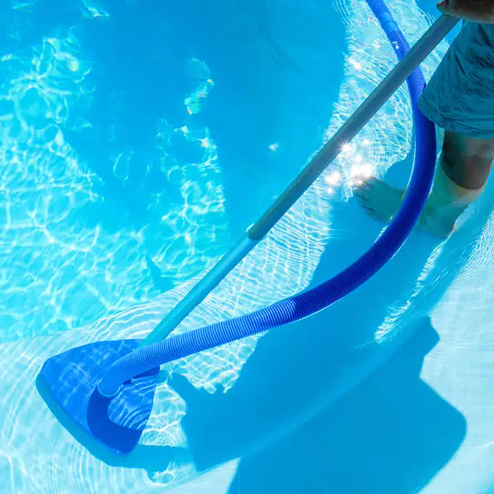Person vacuuming a swimming pool with a blue hose and manual pool cleaner on a sunny day